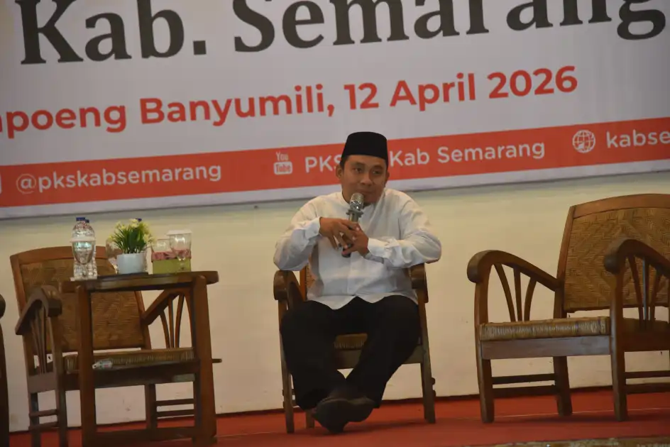 Seorang pria mengenakan baju koko putih dan peci hitam duduk di kursi sambil memegang mikrofon, di panggung dengan spanduk bertuliskan “Kampoeng Banyumili, 12 April 2026” di belakangnya; di sampingnya terdapat meja kayu dengan gelas dan pot bunga.